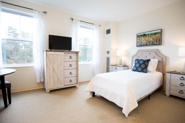 A bright and cozy bedroom with a white bed, dresser with a TV, and large windows accentuated by light curtains.