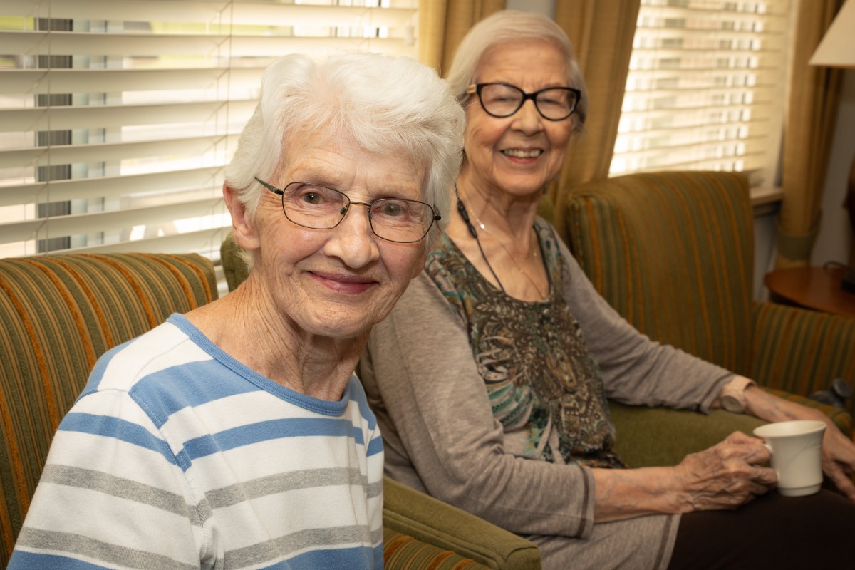 Two elderly women smiling and sitting indoors.