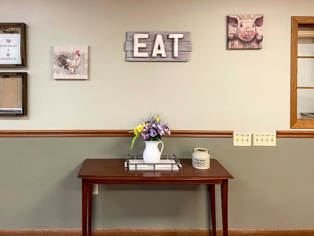 A cozy kitchen setup featuring a wooden table with a ceramic pitcher of flowers, surrounded by playful pig-themed wall art and a rustic 'EAT' sign.
