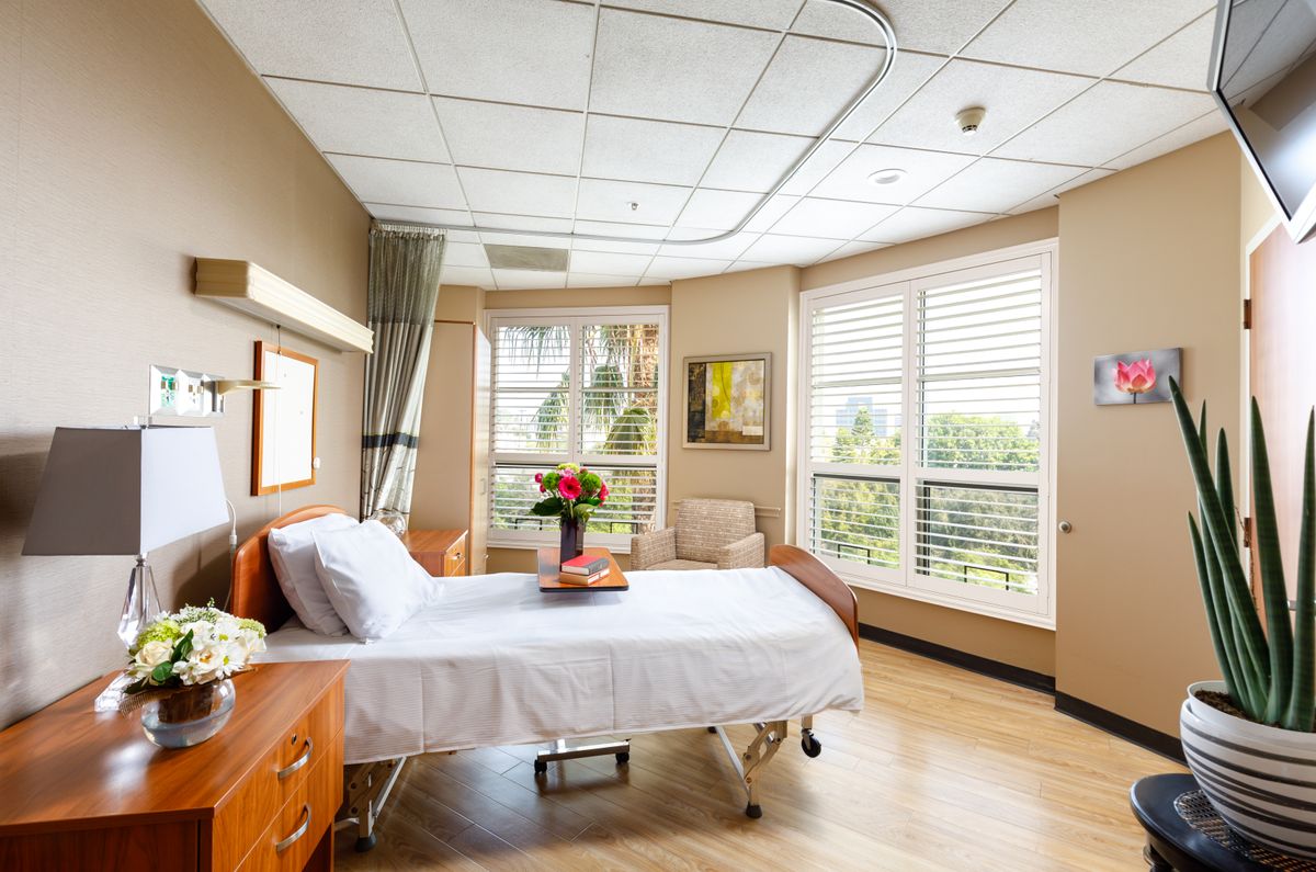 A bright and well-decorated hospital room with a single patient bed, an armchair, and a vase of flowers by the window.