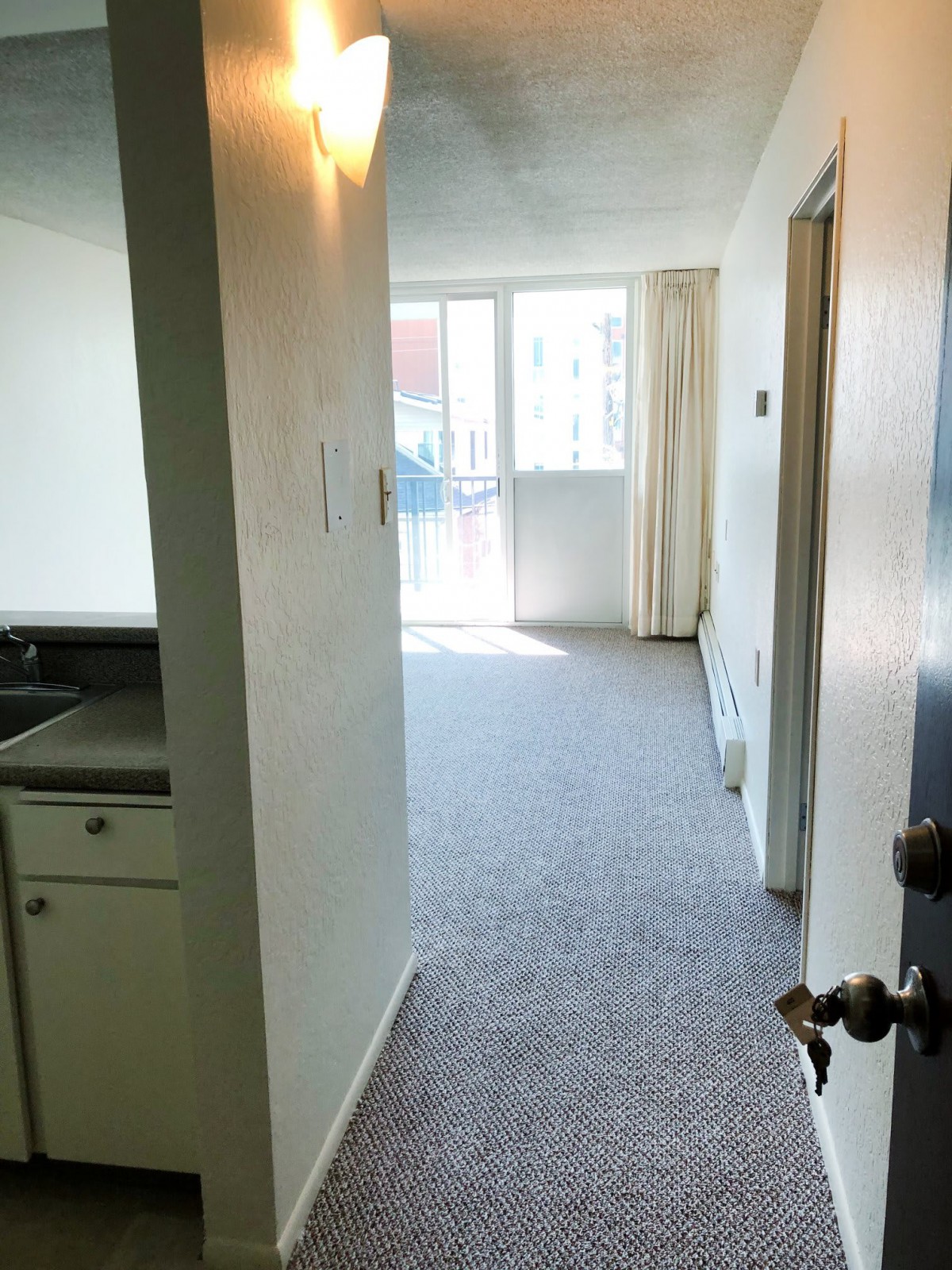 A view of an apartment hallway with carpeted flooring and natural light coming from a window at the end.
