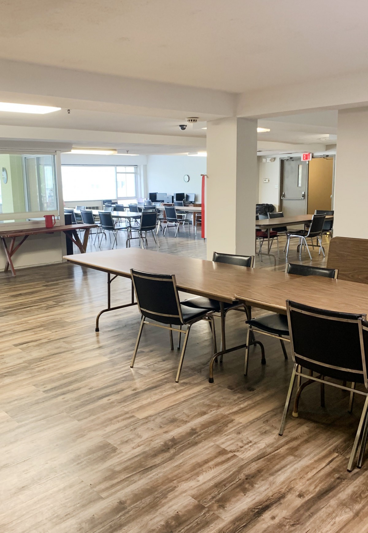 A spacious meeting room with several tables and chairs, featuring large windows providing natural light.