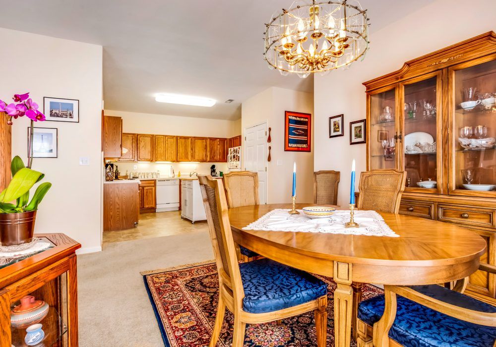 A cozy dining area with a wooden table and chairs, accented by a chandelier, adjacent to an open kitchen.