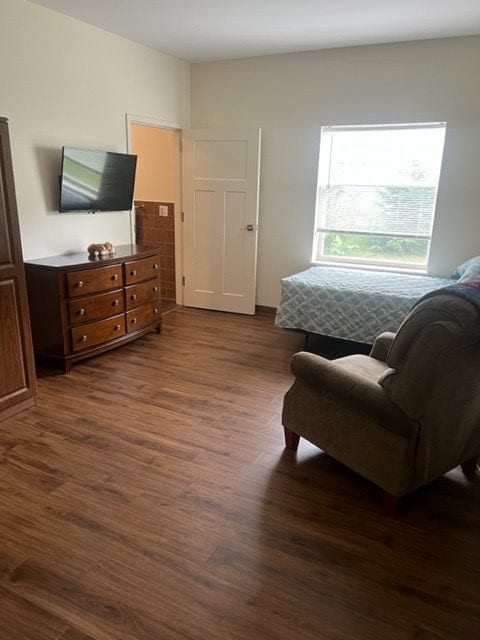A cozy bedroom with wooden flooring, featuring a bed, an armchair, a dresser, and a wall-mounted TV.