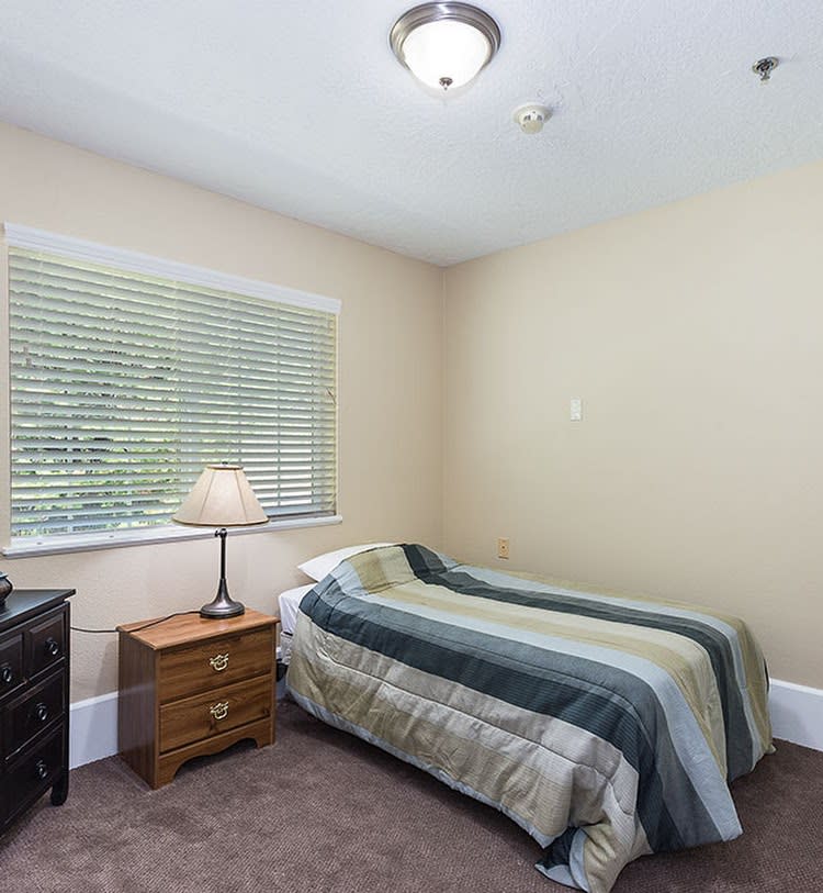 A small bedroom with a single bed, nightstand, and lamp near a window with blinds.