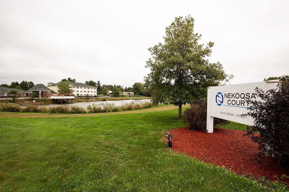 A scenic view of a park featuring a sign, a tree, grassy area, and a lake with buildings in the background.