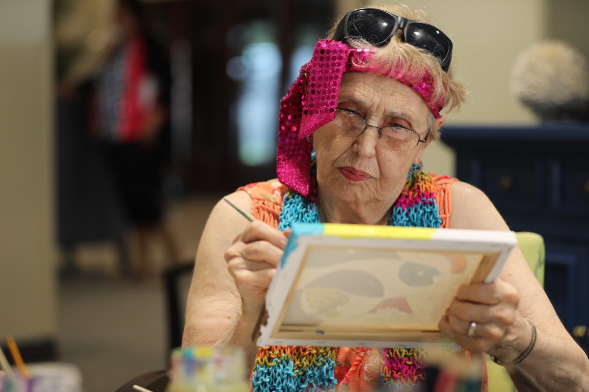 An elderly woman focused on painting, using vibrant colors and art supplies indoors.