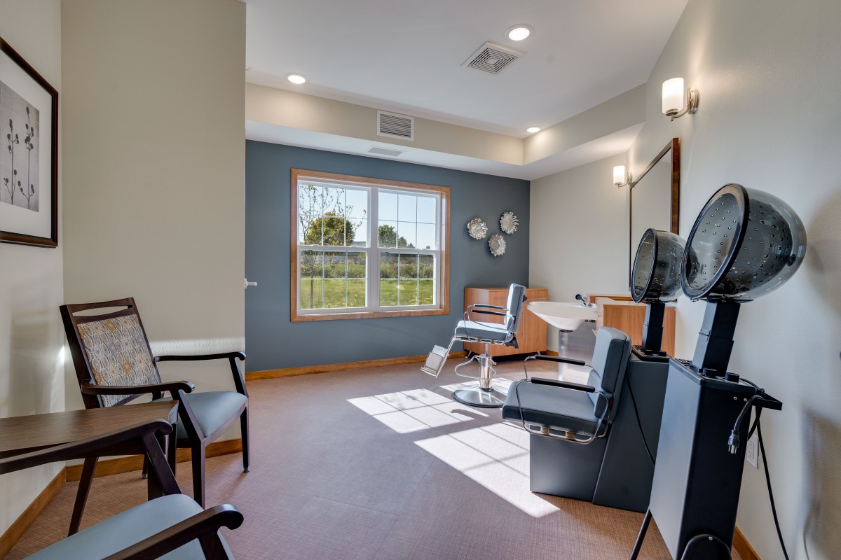 A well-lit salon room with a barber chair, large window, mirror, and seating area.