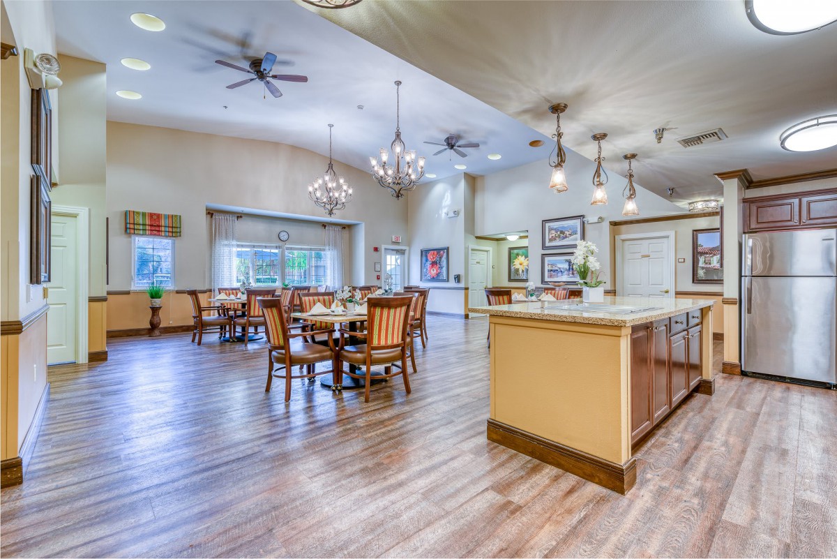 A spacious, modern dining area with wood flooring, elegant chandeliers, and ceiling fans, featuring tables and chairs arranged for comfort.