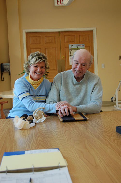 An elderly couple smiling and holding hands at a table indoors.