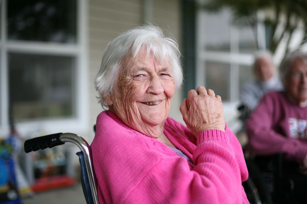 An elderly woman in a pink sweater smiles warmly while seated outdoors.