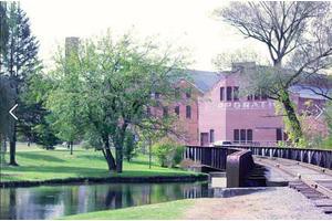 A serene landscape featuring a brick building, a small bridge over water, and lush green trees.