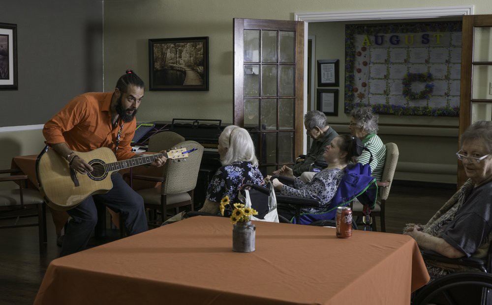 A musician plays the guitar for a group of seniors in a nursing home setting.
