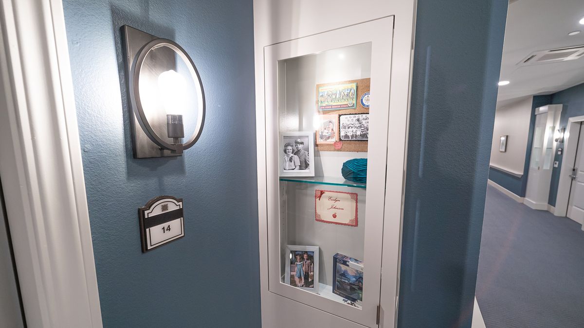A cozy corner featuring a wall-mounted glass cabinet displaying family photos and memorabilia, illuminated by a nearby sconce on a blue-painted wall.