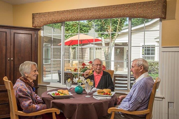 Three seniors enjoying a meal together by a large window overlooking a patio.