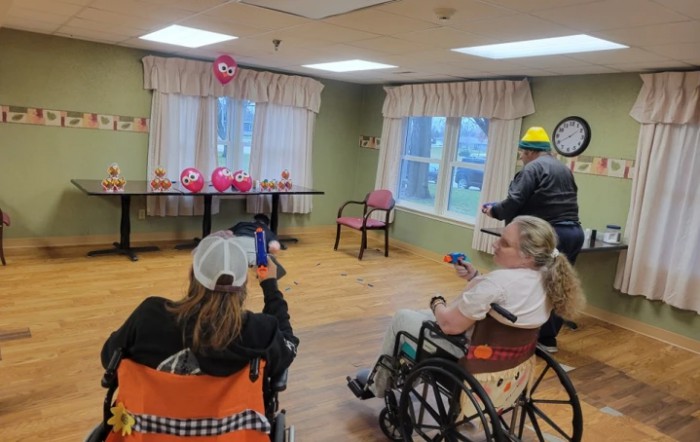 A group of people in wheelchairs participating in an indoor activity, with balloons and decorations visible in the background.