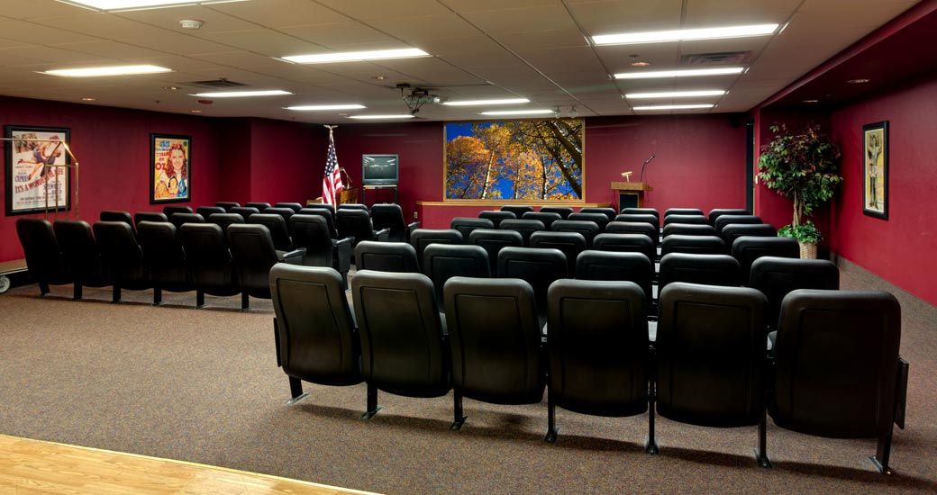An empty conference room with rows of chairs facing a podium and projector screen.