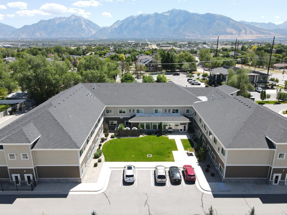 Aerial view of a residential building with a green lawn and parking lot, set against a backdrop of mountains.