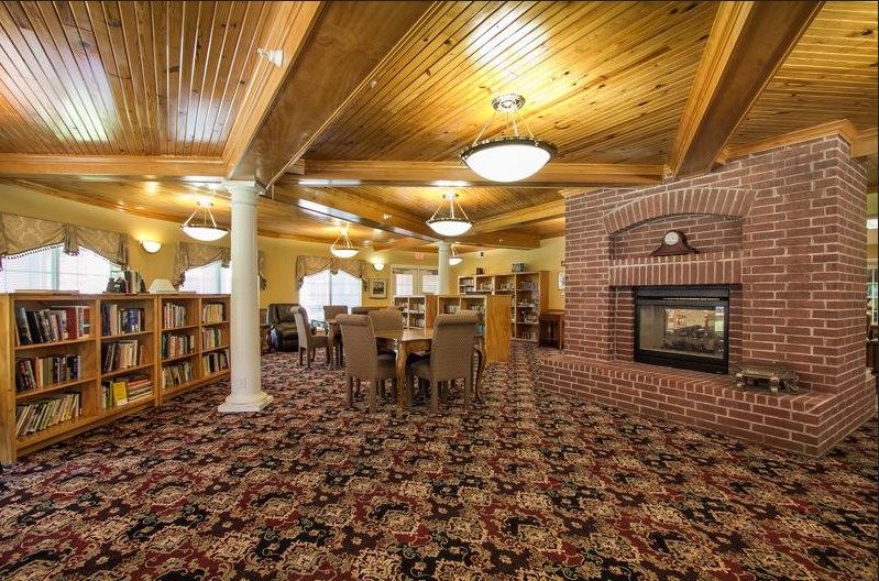A cozy library interior featuring a brick fireplace, wooden ceiling, and carpeted floor with tables and bookshelves.