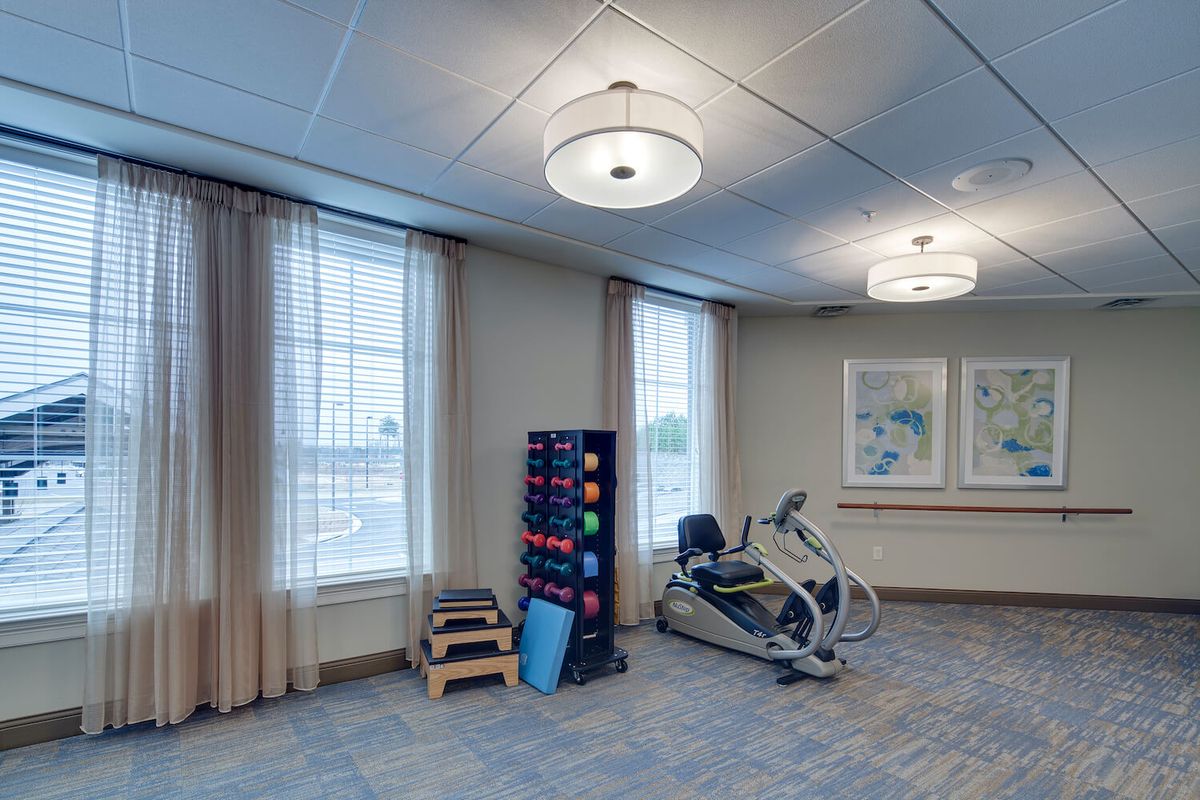 A well-lit exercise room with fitness equipment, including a chair exercise machine and stacked mats.