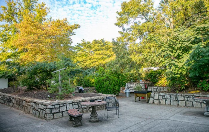 A tranquil garden with a stone patio and outdoor seating, surrounded by lush greenery.