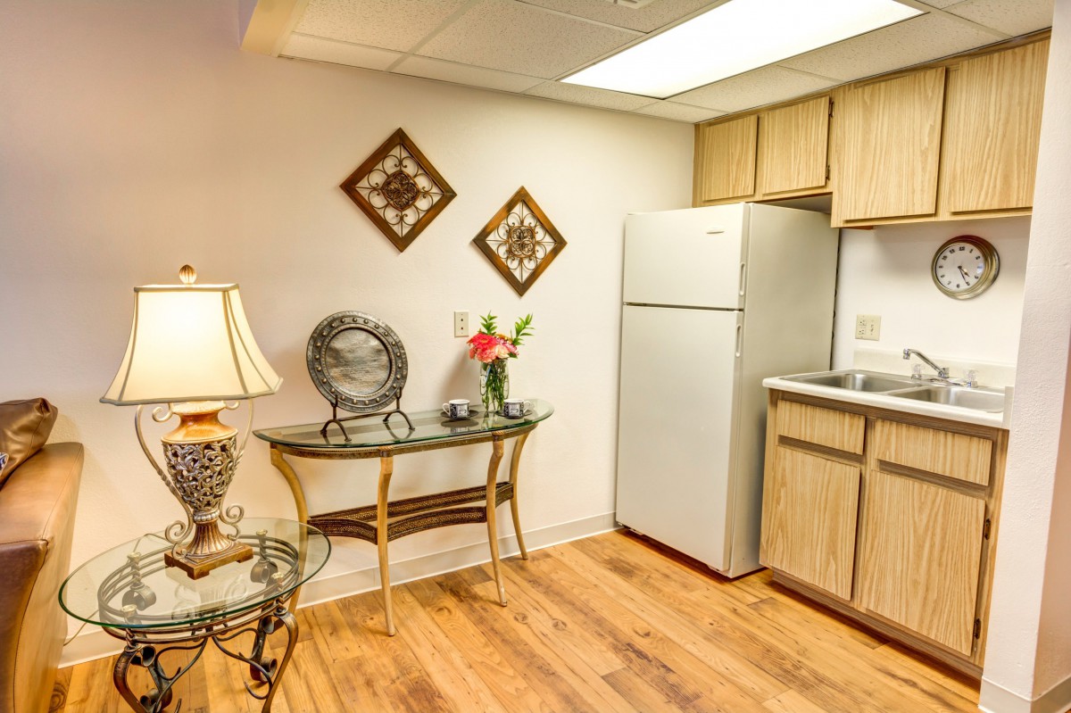 A cozy kitchen area with wooden floors, a fridge, and decorative elements such as a table with flowers, a lamp, and wall art.