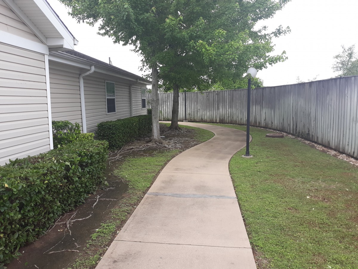 A winding sidewalk beside a building, bordered by grass and bushes with a tree and fence in the background.