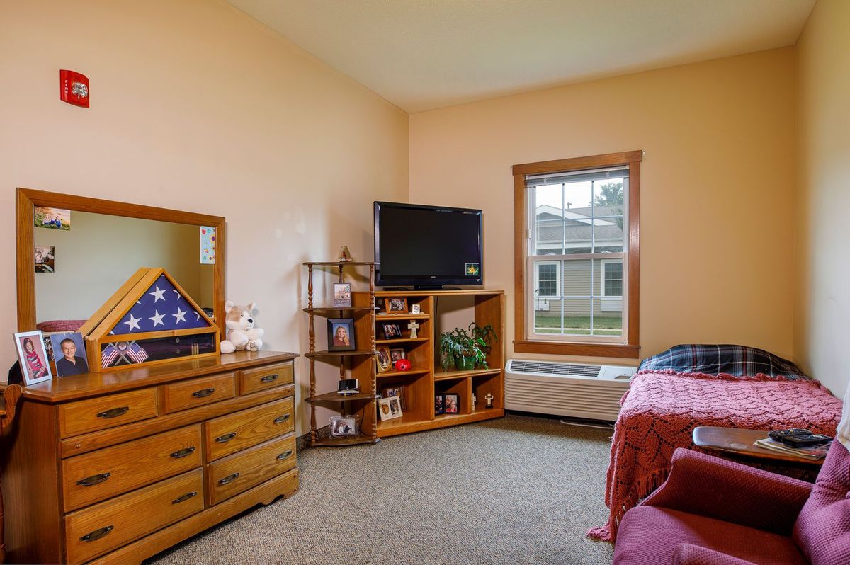 A cozy bedroom with a neatly arranged dresser, television, and a window overlooking the outside.
