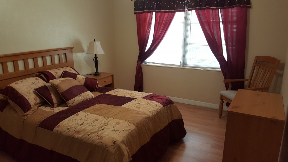 A cozy bedroom with a neatly made bed, bedside lamp, and large window featuring red curtains.