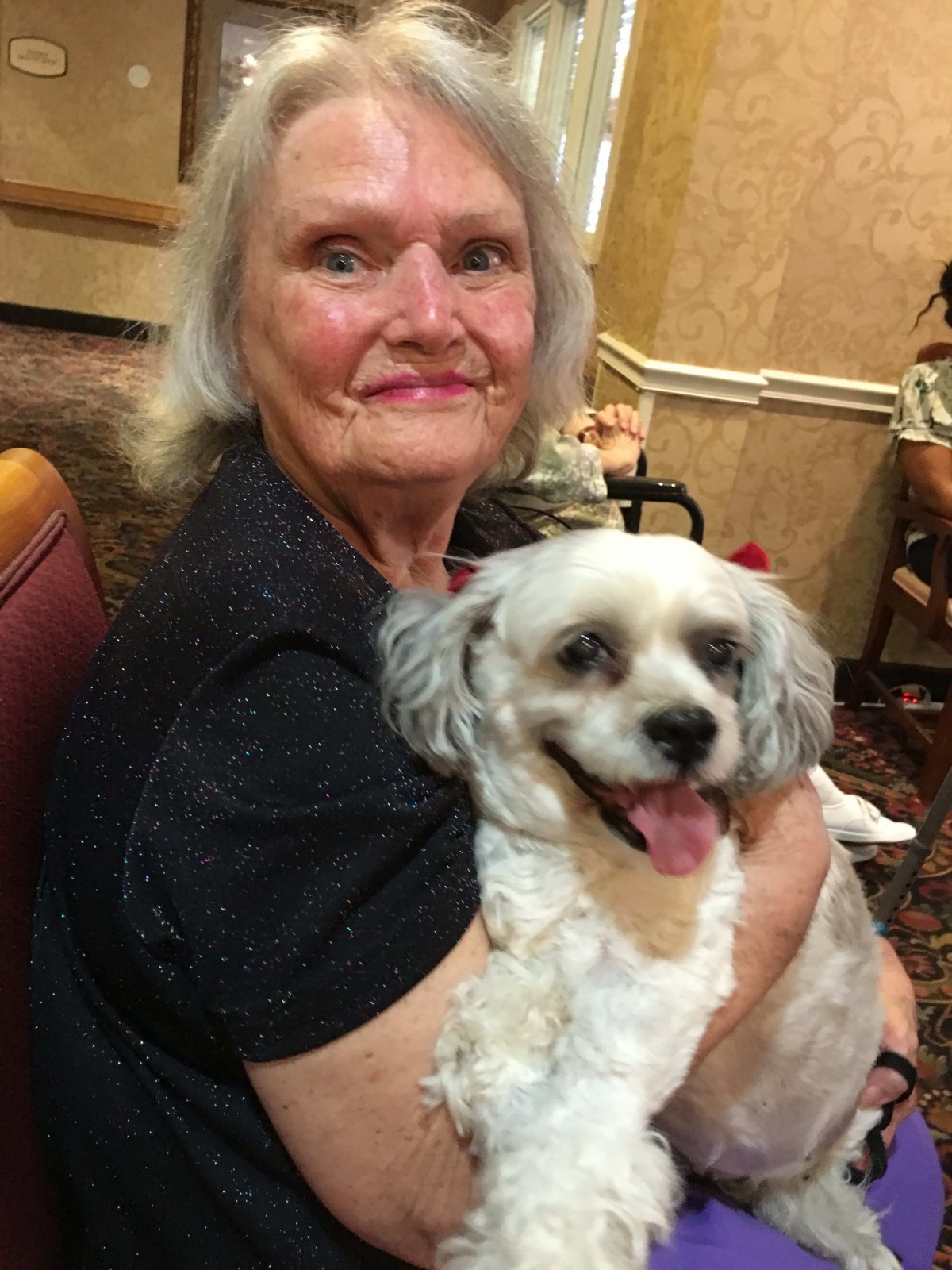 An elderly woman smiling while holding a happy dog indoors.