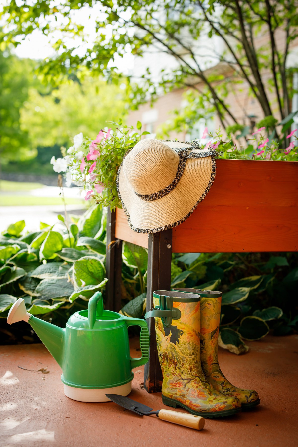A sun hat and a green watering can next to a raised planter filled with blooming flowers.