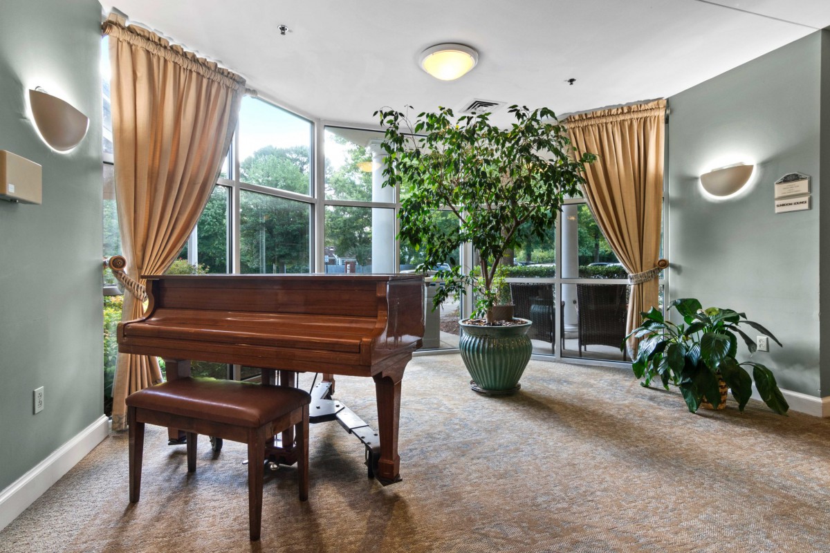 A cozy music room with a grand piano, surrounded by large windows and indoor plants.