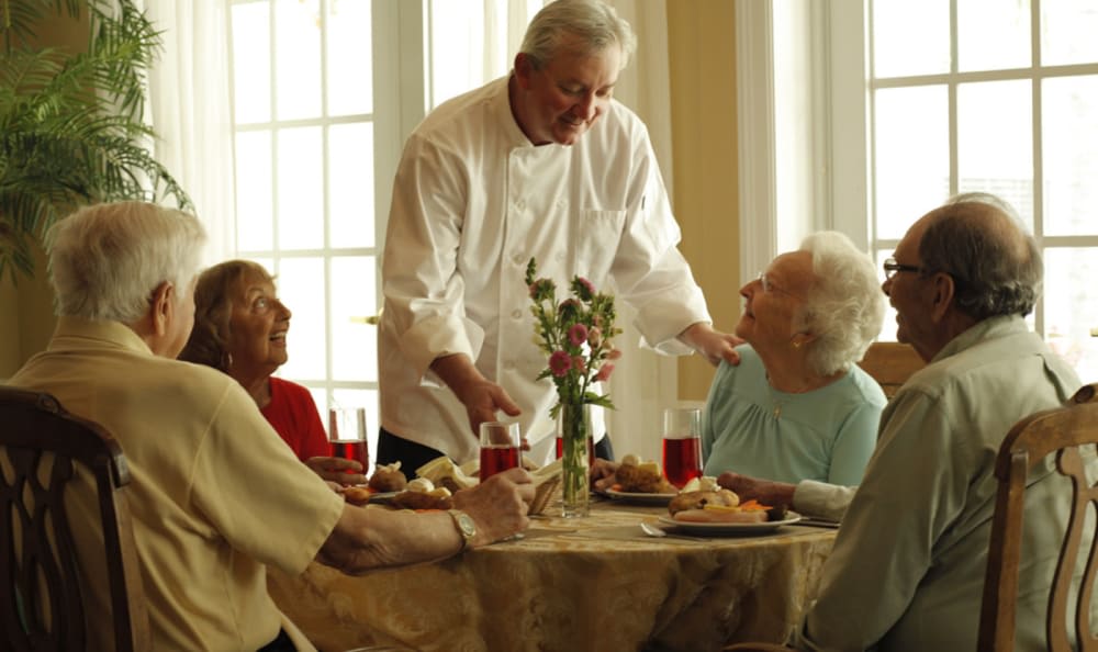 An elderly group enjoys a meal served by a waiter in a bright dining setting.