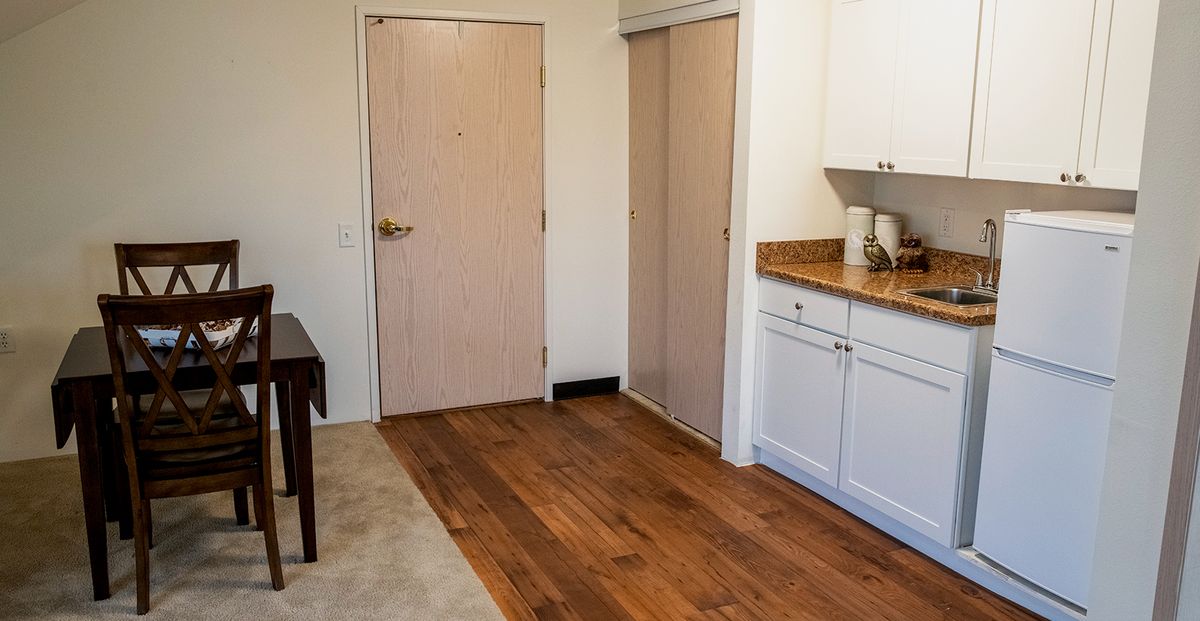 A small kitchen area with wooden flooring, white cabinets, and a countertop adjacent to a doorway.