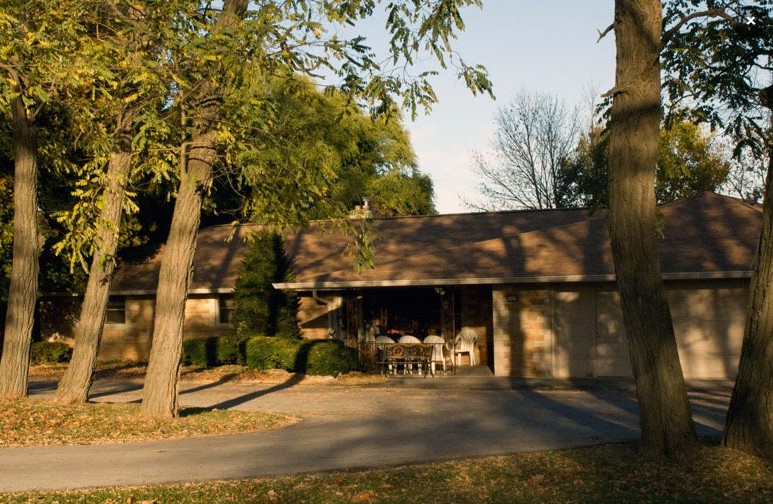 A suburban house surrounded by trees with a driveway in the foreground, bathed in sunlight.