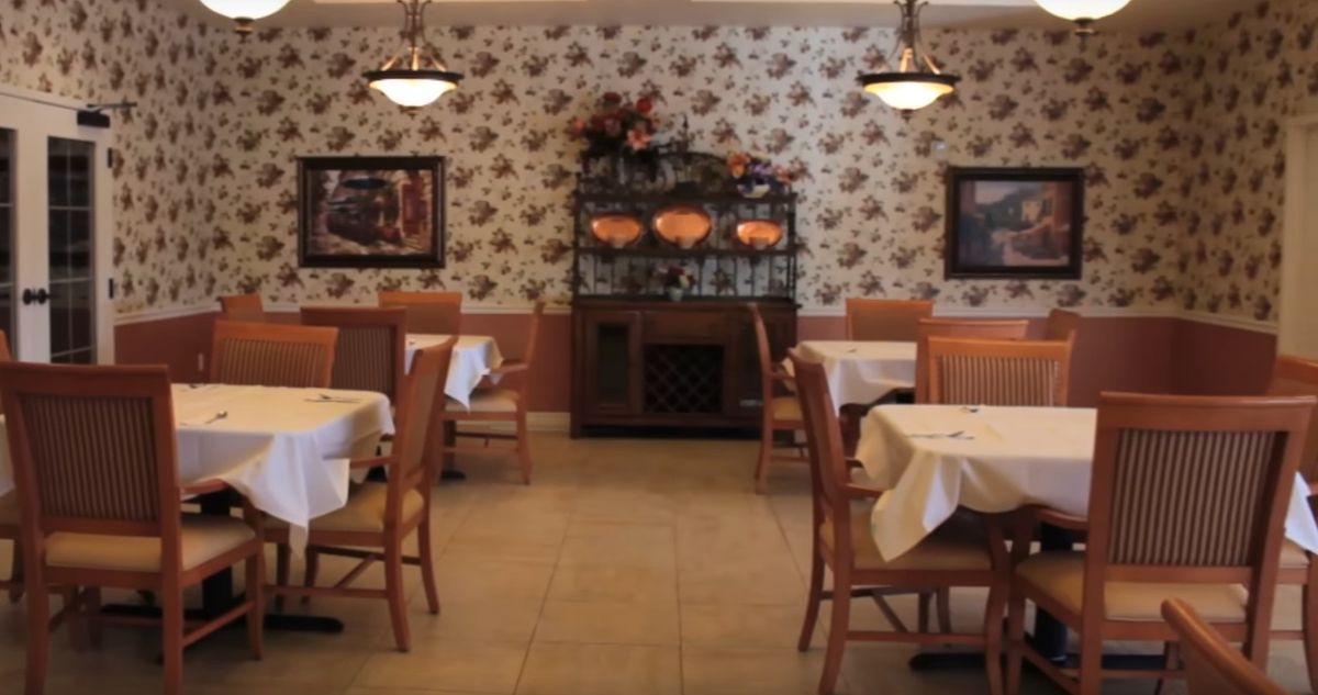 A cozy restaurant interior with tables and chairs arranged neatly, decorated with floral wallpaper and dim lighting.