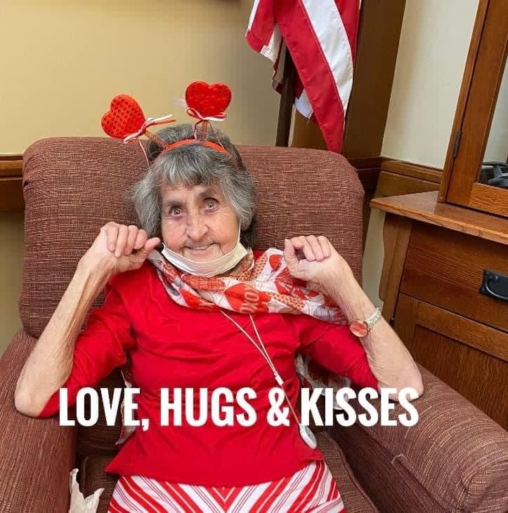 An elderly woman wearing a red outfit and heart-themed accessories, sitting in a chair with an American flag in the background, spreading love, hugs, and kisses.