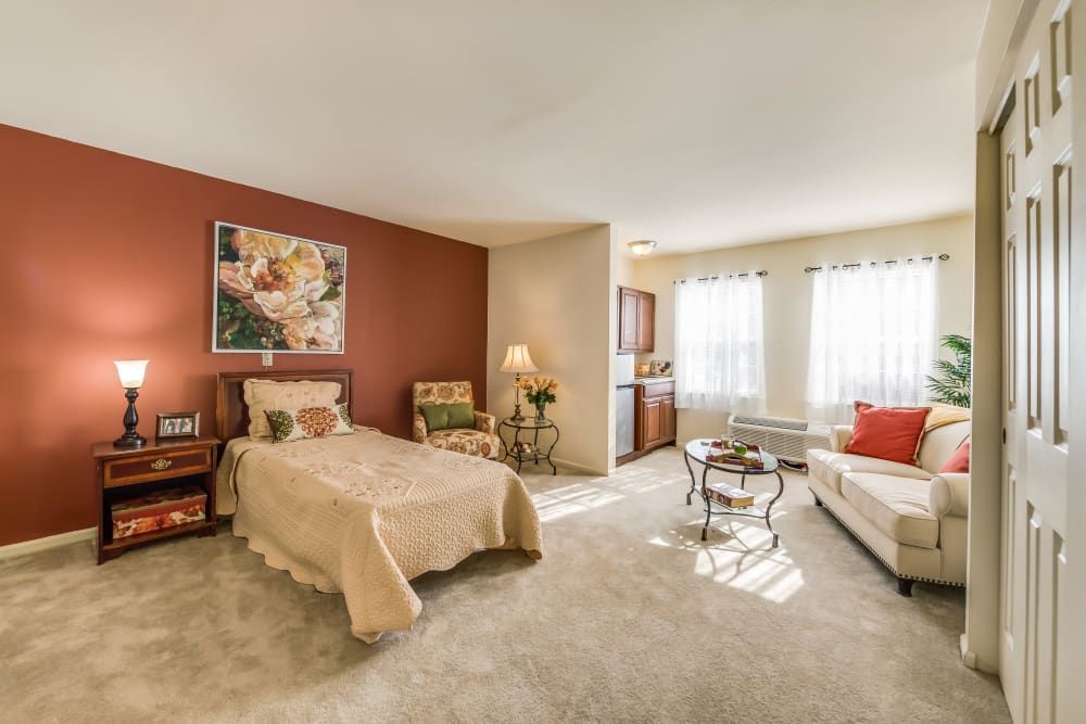 A cozy bedroom with a bed, chairs, and a small kitchenette, bathed in natural light.