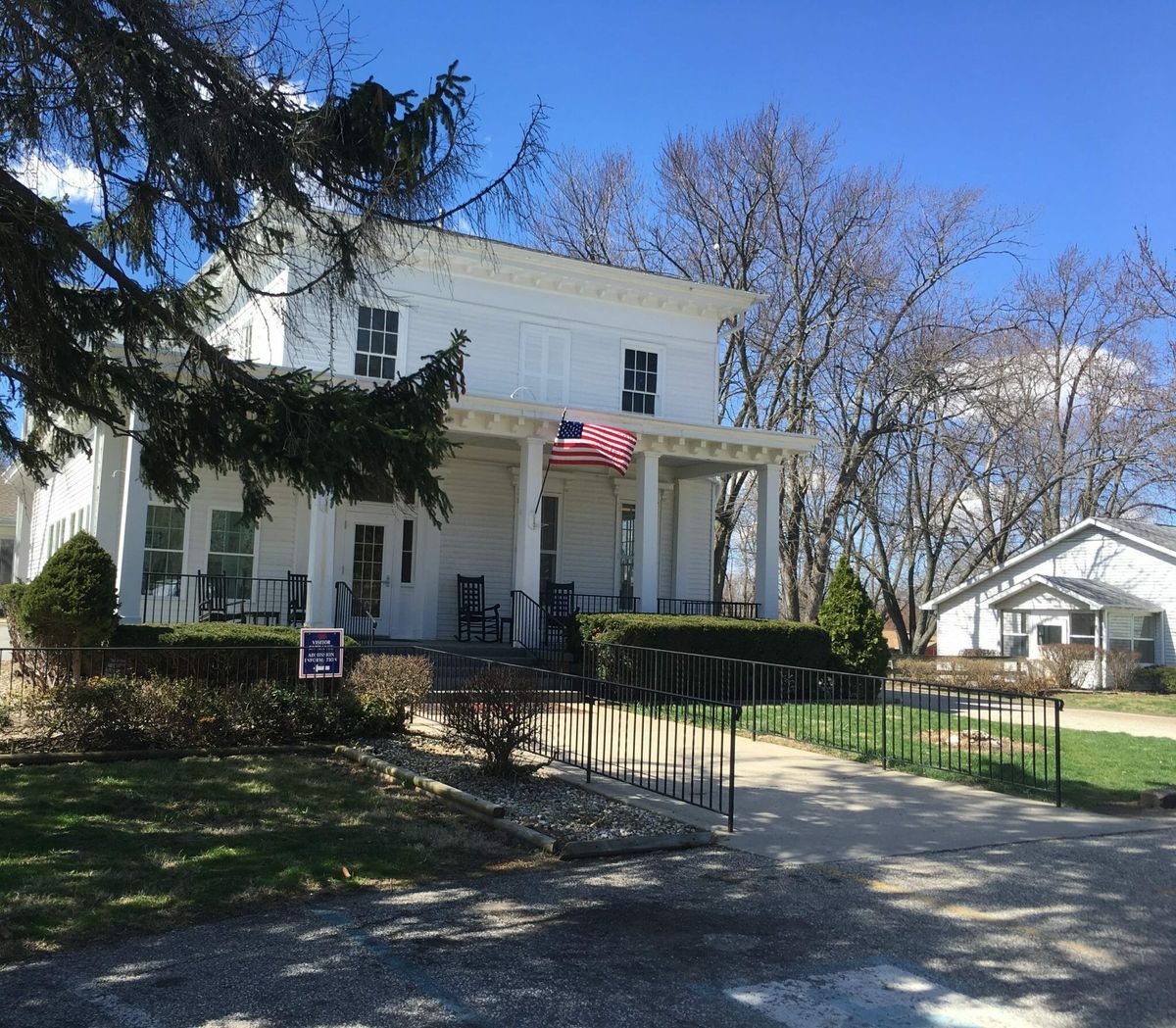 A charming two-story house with a front porch and an American flag, surrounded by trees on a sunny day.