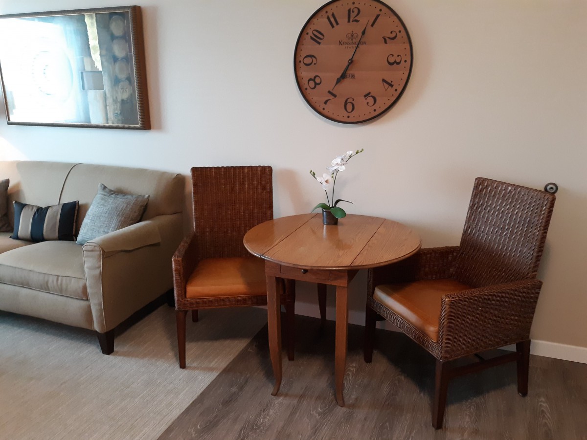 A cozy living room corner featuring two chairs, a round wooden table with a decorative flower arrangement, and a wall clock above.