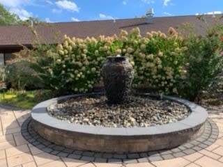 A garden fountain with an urn centerpiece surrounded by plants and a paved patio.