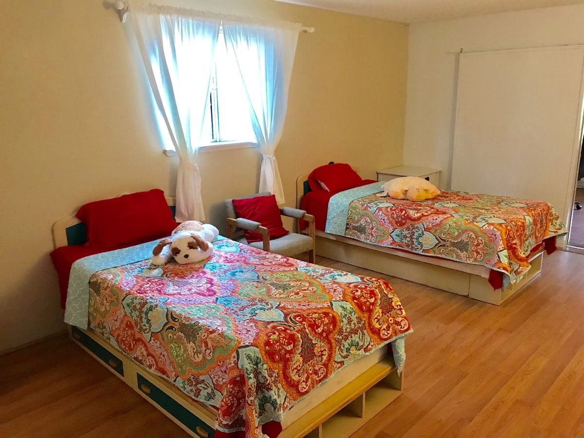 A cozy bedroom with two twin beds adorned with colorful bedding and stuffed animals, featuring a wooden floor and a window with white curtains.