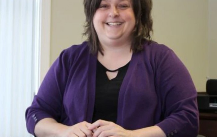 A woman is smiling while sitting indoors, wearing a purple sweater.