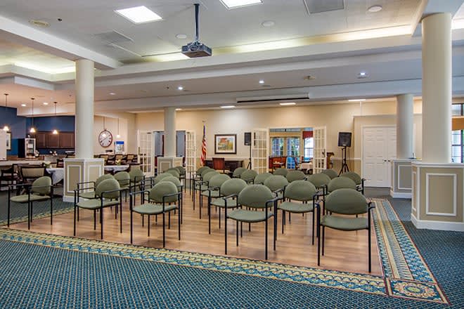 An indoor meeting room with rows of chairs facing a stage area and a projector hanging from the ceiling.