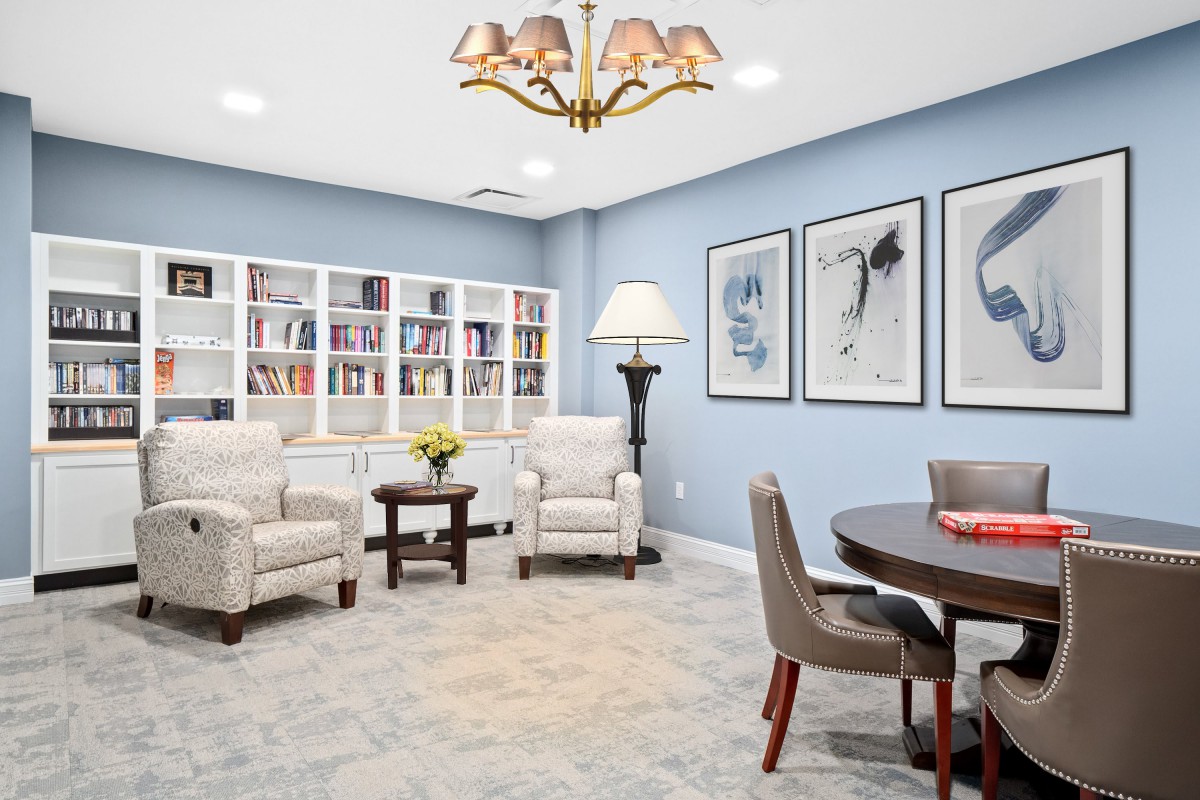A cozy living room with bookshelves, armchairs, and a round table, featuring elegant artwork on the walls.