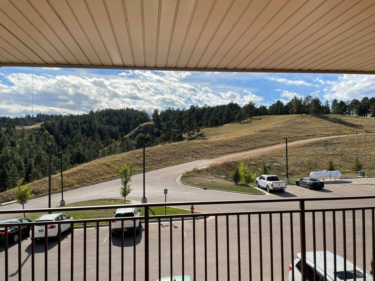 View from a balcony overlooking a winding road with cars, surrounded by trees and a partly cloudy sky.