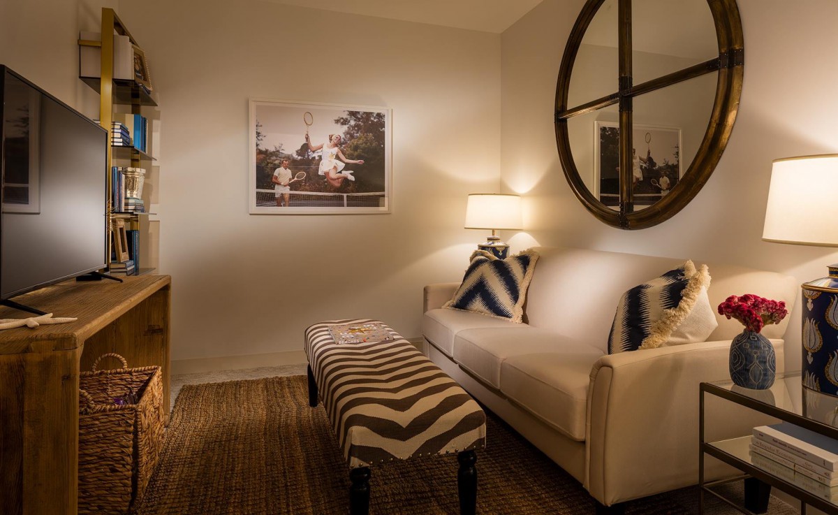 A cozy living room with a white sofa, decorative pillows, a bench with a zigzag pattern, a lamp on the side table, and wall art illuminated by warm lighting.