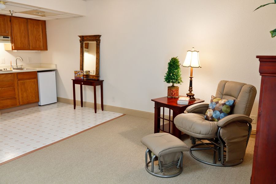 A cozy living room area featuring a comfortable chair with an ottoman, a side table with a lamp and potted plant, and a small mirrored table in the corner.