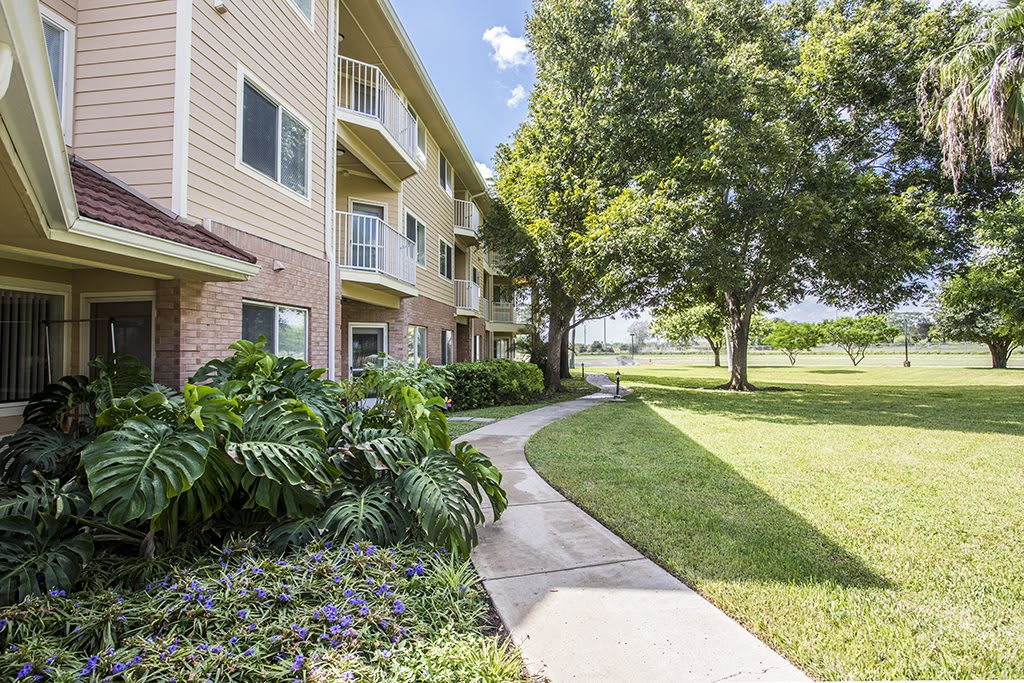 A sunny day outside an apartment building with a well-maintained pathway and lush landscaping.