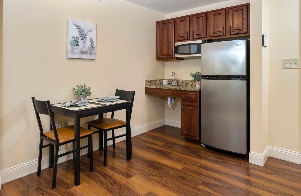 A compact kitchen space with a dining table and chairs, featuring wooden cabinets, a stainless steel refrigerator, and a microwave.